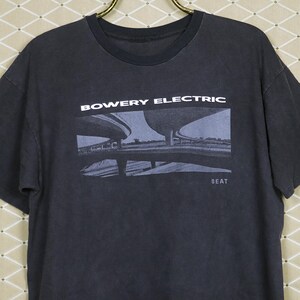 May include: Dark gray t-shirt with the words "BOWERY ELECTRIC" above a grayscale image of a bridge. The word "BEAT" is below the image. The shirt is hanging on a gold-colored hanger.