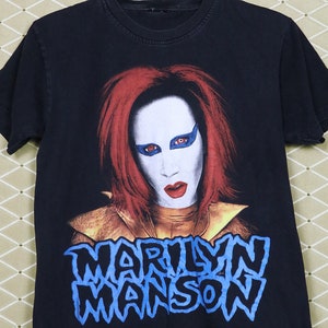 Op de afbeelding: Zwart T-shirt met een grafische print van Marilyn Manson. De afbeelding toont een close-up van Manson met rood haar en een wit masker met blauwe ogen. De tekst "Marilyn Manson" is in blauw onder de afbeelding gedrukt.