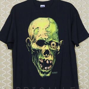 May include: Black t-shirt featuring a green and yellow zombie skull graphic. The design includes details like exposed teeth and a single visible eye. The shirt is hanging on a gold-colored hook.