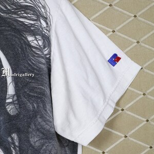 May include: White t-shirt featuring a black and white graphic print of a woman's face. The shirt has a small blue and red logo on the right sleeve. The text "Madrigalgallery" is visible on the shirt.