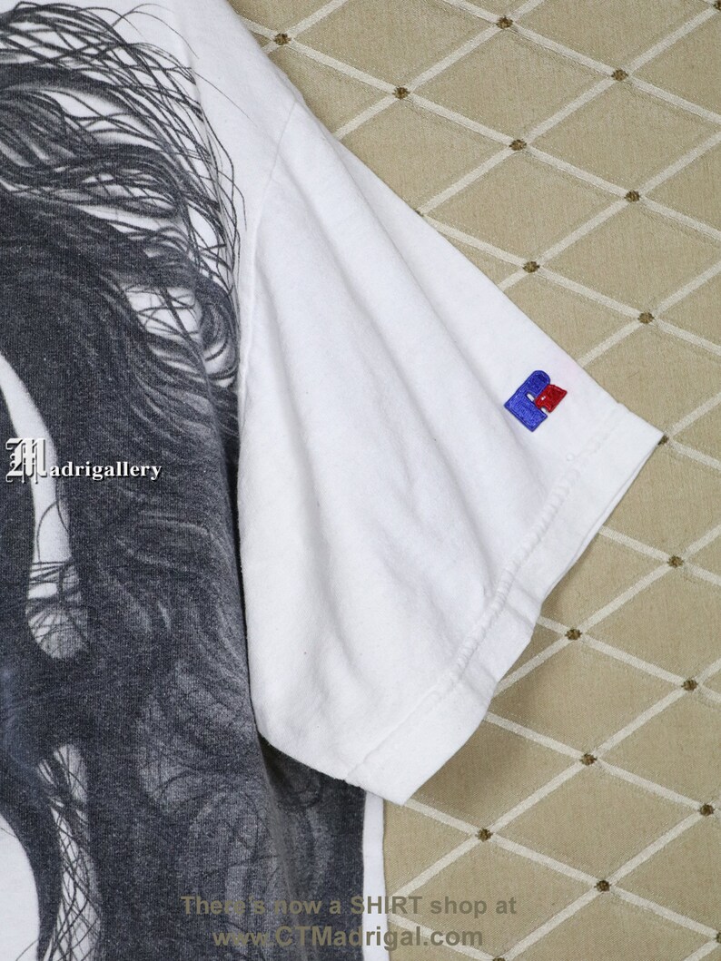 May include: White t-shirt featuring a black and white graphic print of a woman's face. The shirt has a small blue and red logo on the right sleeve. The text "Madrigalgallery" is visible on the shirt.