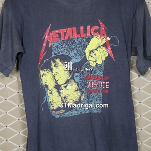 May include: A vintage blue t-shirt with a graphic of the band Metallica. The graphic features a hand holding a microphone with the band's name in red and the words "Hammer of Justice" in white. The band's website, "CTMadrigal.com", is printed at the bottom of the graphic.