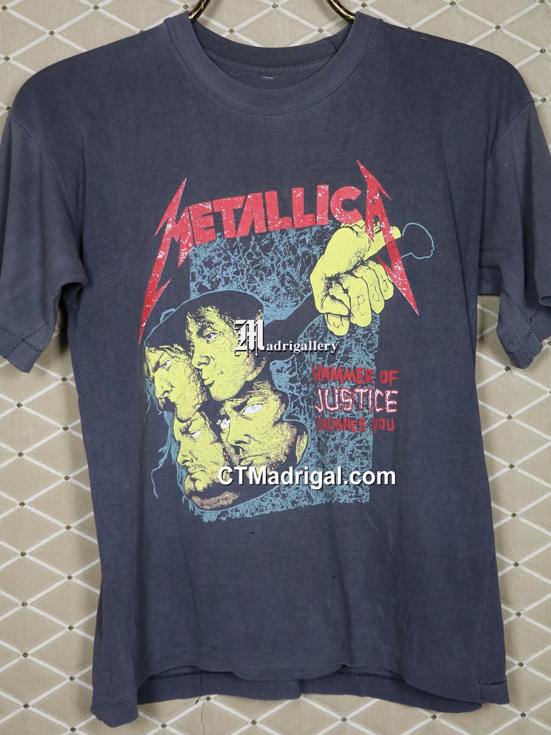 May include: A vintage blue t-shirt with a graphic of the band Metallica. The graphic features a hand holding a microphone with the band's name in red and the words "Hammer of Justice" in white. The band's website, "CTMadrigal.com", is printed at the bottom of the graphic.