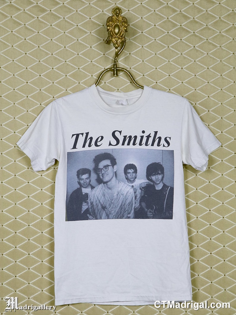 K&ouml;nnte beinhalten: Wei&szlig;es T-Shirt mit dem Bandnamen "The Smiths" in Schwarz &uuml;ber einem Schwarz-Wei&szlig;-Bild der Band. Das T-Shirt h&auml;ngt an einem goldfarbenen Kleiderb&uuml;gel. Der Hintergrund ist eine gemusterte, hellfarbige Tapete.
