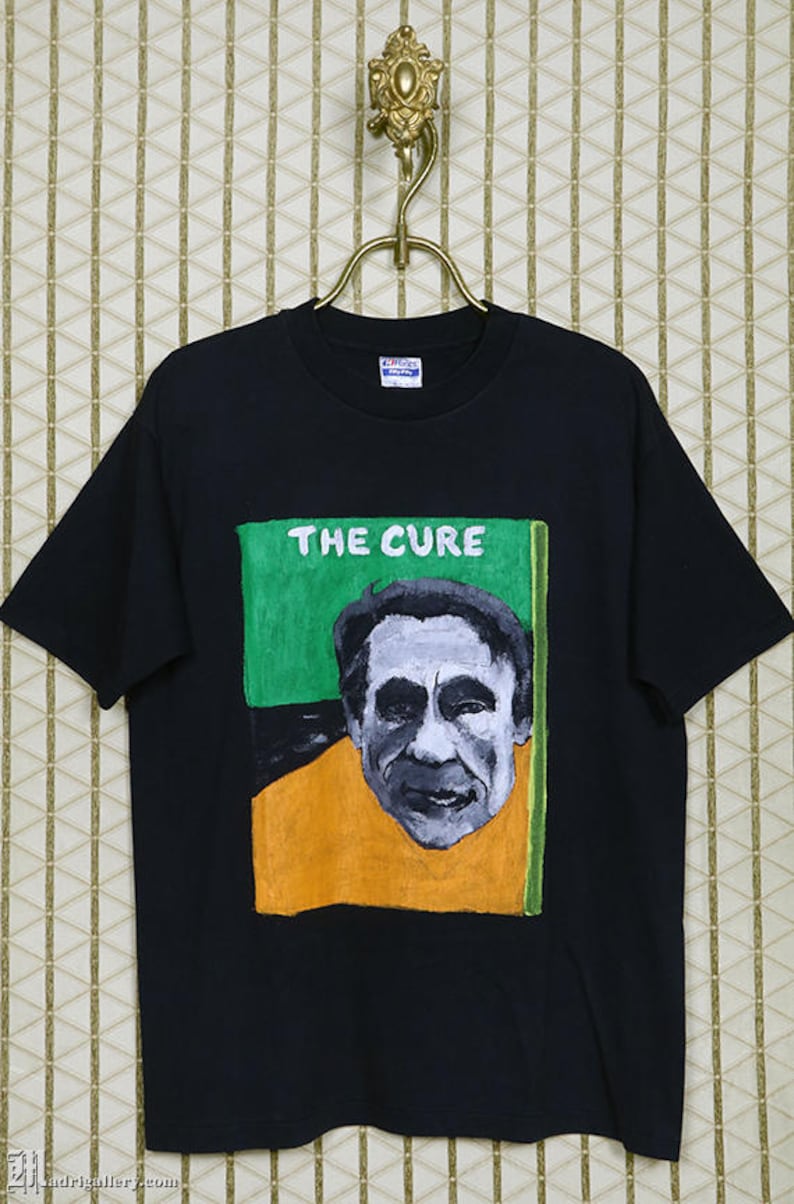 May include: Black t-shirt with a graphic print of Robert Smith, the lead singer of The Cure, in a yellow shirt and green background. The text "THE CURE" is printed in white above the image.