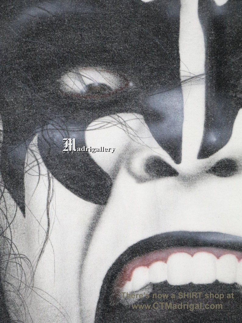May include: A close-up black and white image of a person's face with a fierce expression. The person has long black hair and is wearing black makeup around the eyes. The mouth is open wide, revealing white teeth. The image is slightly grainy and has a vintage feel. There's now a SHIRT shop at www.CTMadrigal.com