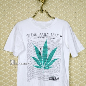 Op de afbeelding: Wit T-shirt met een krantenstijl grafische afbeelding met een groot groen cannabisblad. De tekst "THE DAILY LEAF CANNABIS HISTORY" staat bovenaan. Het shirt heeft ook de tekst "There's now a SHIRT shop at www.CTMadrigal.com".