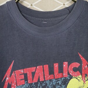 May include: A dark gray t-shirt with a red and white distressed "Metallica" logo printed on the front. The logo is set against a black and blue background with a yellow hand reaching out from the bottom.