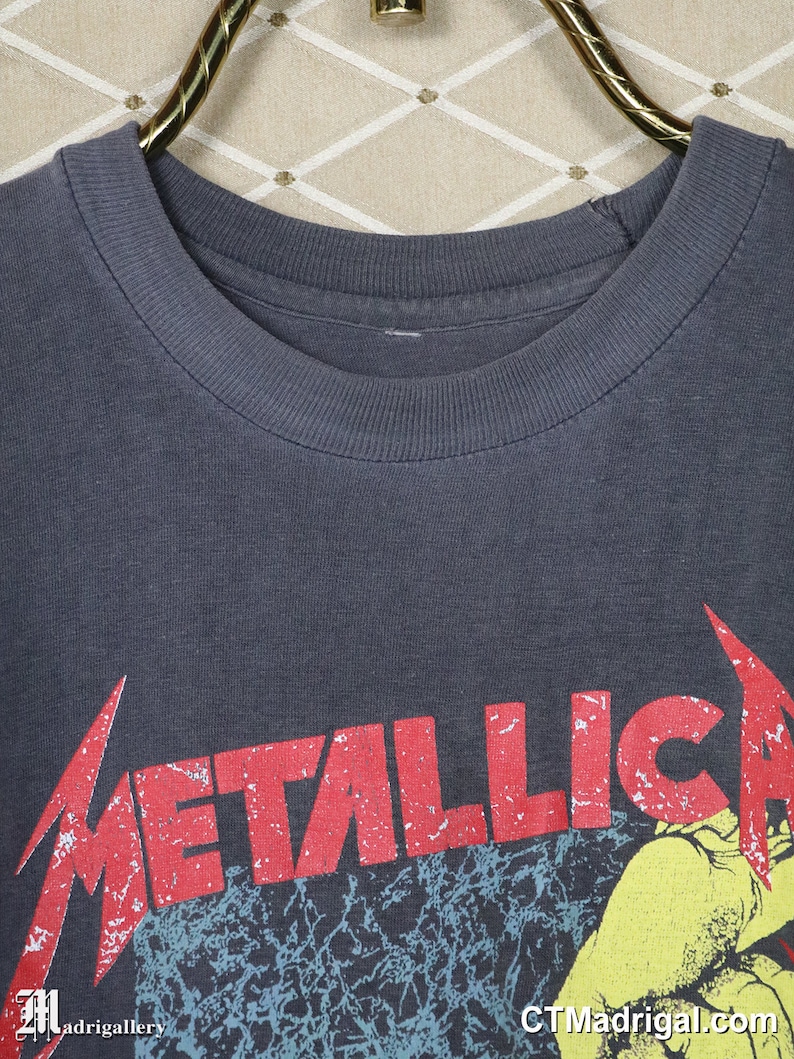 May include: A dark gray t-shirt with a red and white distressed "Metallica" logo printed on the front. The logo is set against a black and blue background with a yellow hand reaching out from the bottom.