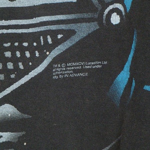 Op de afbeelding: Zwart T-shirt met een blauwe en witte afbeelding van Darth Vaders helm. De tekst "TM & &copy; MCMXCVI Lucasfilm Ltd. all rights reserved. Used under authorization. Mfg. by IN ADVANCE" staat gedrukt op de onderkant van het shirt.
