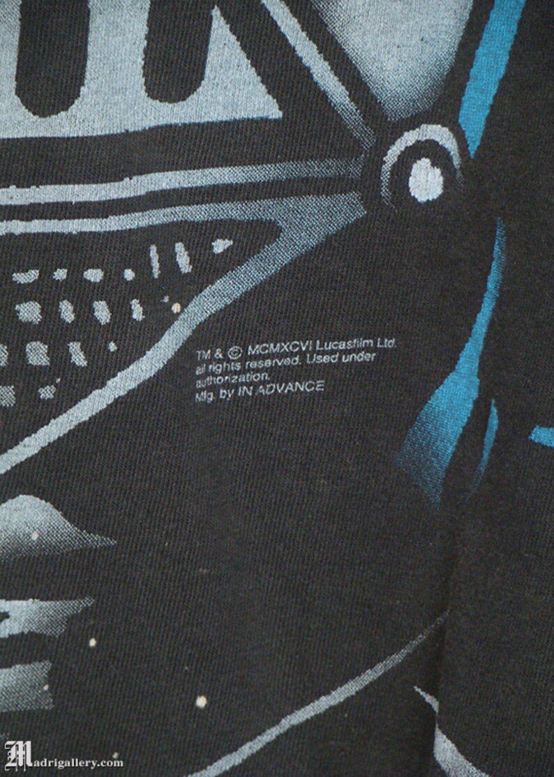 Op de afbeelding: Zwart T-shirt met een blauwe en witte afbeelding van Darth Vaders helm. De tekst "TM & &copy; MCMXCVI Lucasfilm Ltd. all rights reserved. Used under authorization. Mfg. by IN ADVANCE" staat gedrukt op de onderkant van het shirt.