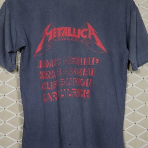 May include: A dark blue t-shirt with a red Metallica logo and the band members' names printed on the back. The names are James Hetfield, Kirk Hammett, Cliff Burton, and Lars Ulrich.