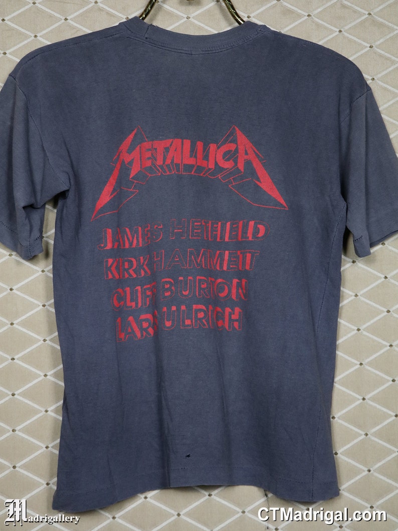 May include: A dark blue t-shirt with a red Metallica logo and the band members' names printed on the back. The names are James Hetfield, Kirk Hammett, Cliff Burton, and Lars Ulrich.