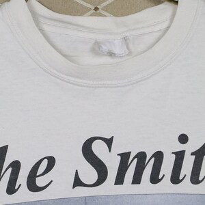 K&ouml;nnte beinhalten: Wei&szlig;es T-Shirt mit dem Bandnamen "The Smiths" in Schwarz aufgedruckt. Das Shirt zeigt ein Schwarz-Wei&szlig;-Bild unter dem Text. Das Shirt h&auml;ngt an einem goldenen Kleiderb&uuml;gel.