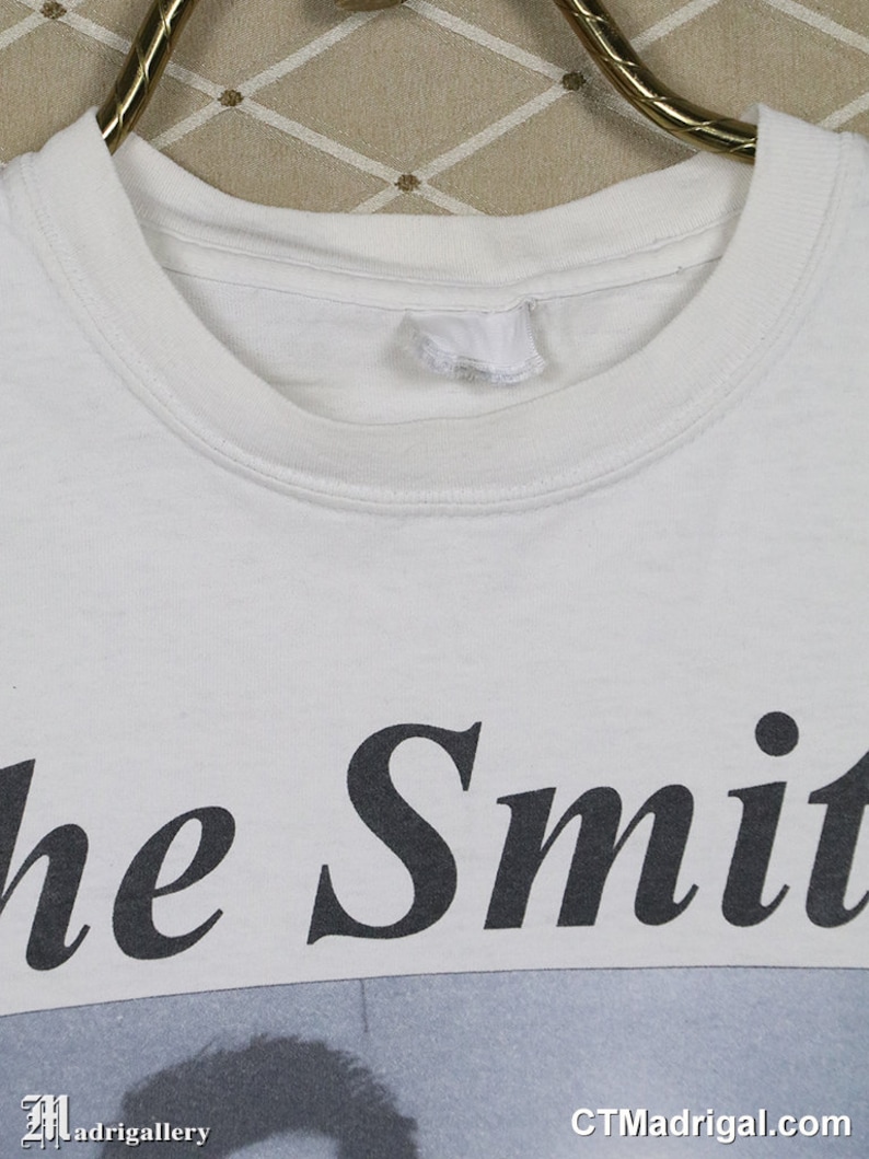 K&ouml;nnte beinhalten: Wei&szlig;es T-Shirt mit dem Bandnamen "The Smiths" in Schwarz aufgedruckt. Das Shirt zeigt ein Schwarz-Wei&szlig;-Bild unter dem Text. Das Shirt h&auml;ngt an einem goldenen Kleiderb&uuml;gel.