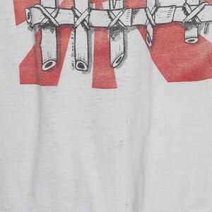 May include: White t-shirt with a red and white graphic print of a face with thorns and a bamboo cage. The text "Gary Scionti '87" is printed on the bottom right corner of the graphic.