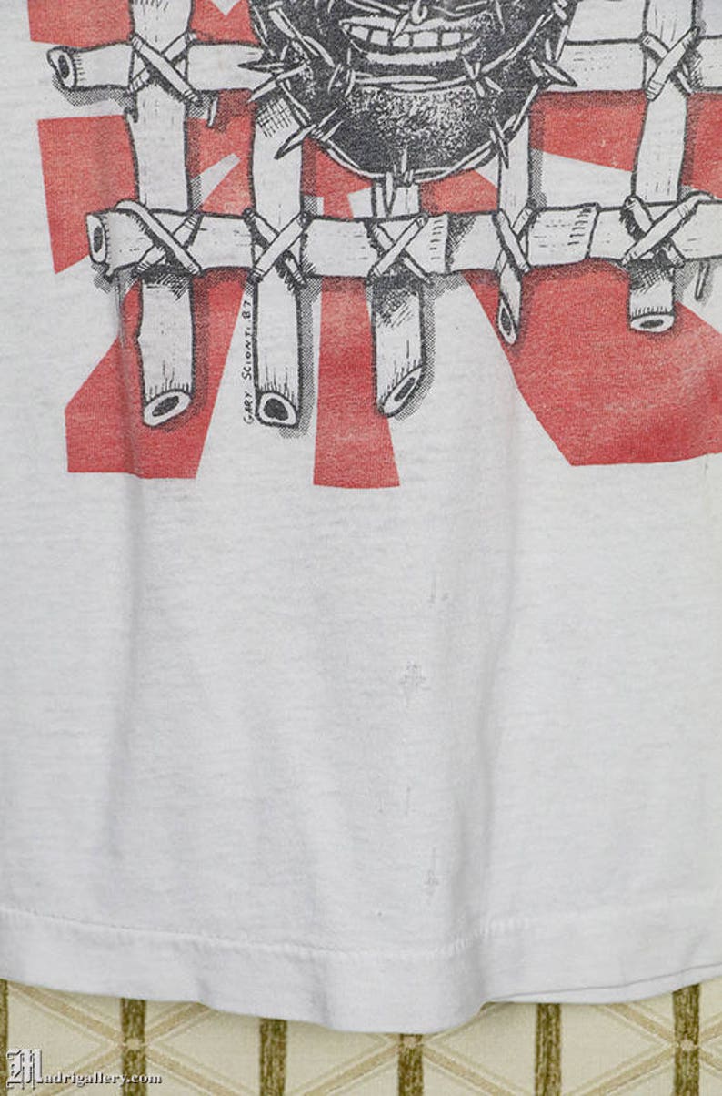 May include: White t-shirt with a red and white graphic print of a face with thorns and a bamboo cage. The text "Gary Scionti '87" is printed on the bottom right corner of the graphic.