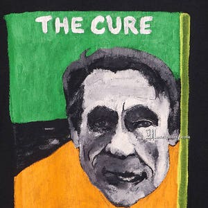 May include: A black t-shirt with a painted image of a man's face with the text "THE CURE" above it. The image is surrounded by a green, orange, and yellow border.