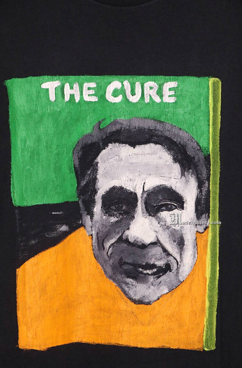 May include: A black t-shirt with a painted image of a man's face with the text "THE CURE" above it. The image is surrounded by a green, orange, and yellow border.