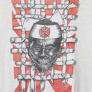 May include: A white t-shirt with a black and white graphic print of a person's face with a barbed wire mouth. The person is wearing a white headband with a red circle and a black symbol inside. The graphic is surrounded by a red and white pattern.