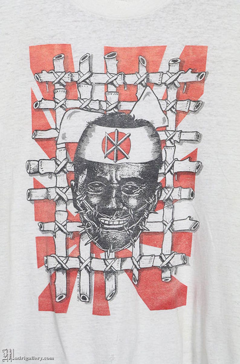 May include: A white t-shirt with a black and white graphic print of a person's face with a barbed wire mouth. The person is wearing a white headband with a red circle and a black symbol inside. The graphic is surrounded by a red and white pattern.