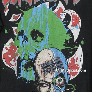 May include: Black t-shirt with a green, blue, and red graphic of a skull with the text "DO NOT PUNCTURE CONTENTS UNDER PRESSURE" on the forehead. The band name "EXODUS" is printed in red at the top of the shirt.