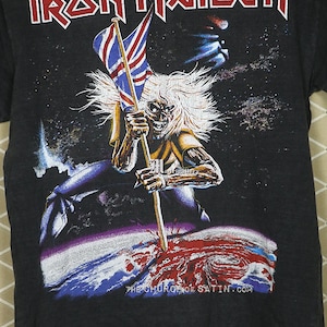 May include: Black t-shirt with a graphic print of a skeleton holding a flag that says "Iron Maiden" in red letters. The skeleton is standing on a planet with a red and blue swirl. The text "The Church of Satin.com" is printed at the bottom of the shirt.