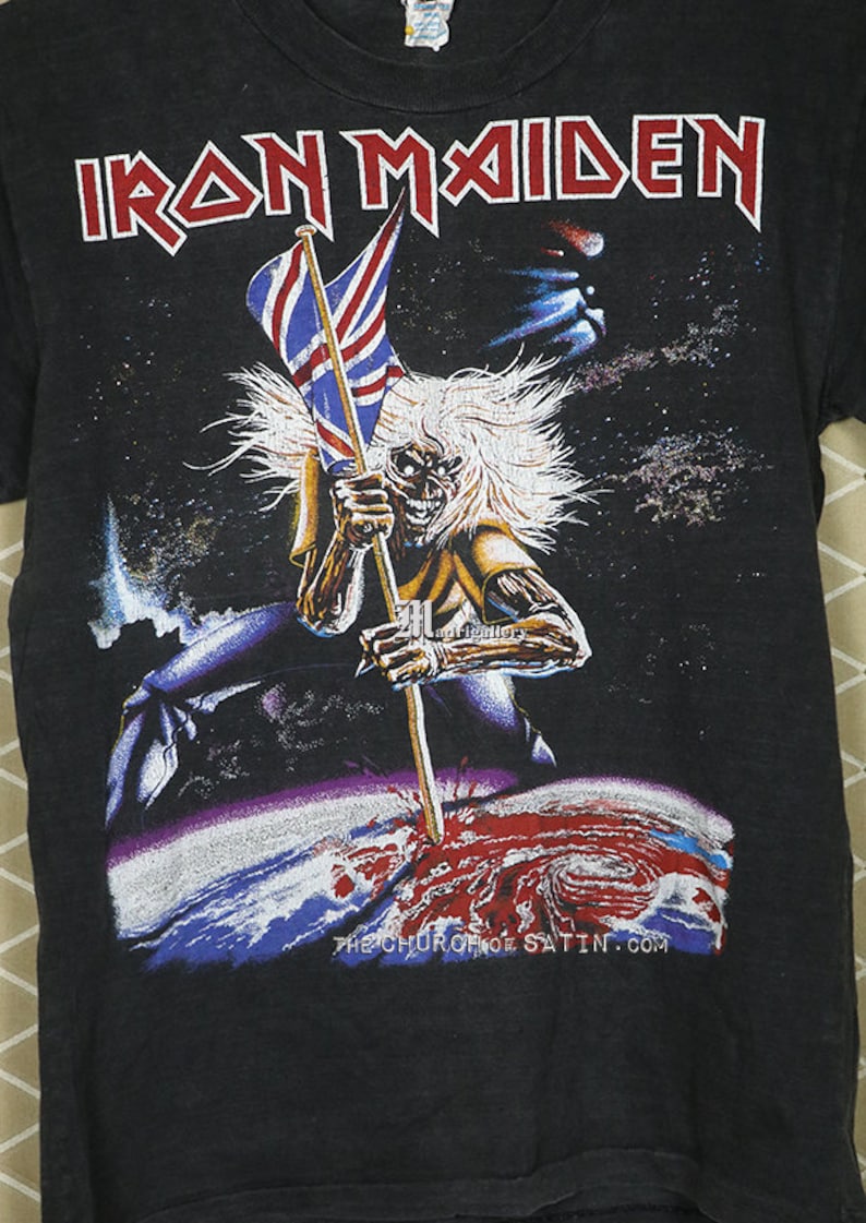 May include: Black t-shirt with a graphic print of a skeleton holding a flag that says "Iron Maiden" in red letters. The skeleton is standing on a planet with a red and blue swirl. The text "The Church of Satin.com" is printed at the bottom of the shirt.