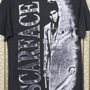 Op de afbeelding: Zwart T-shirt met een witte grafische print van Al Pacino als Tony Montana uit de film Scarface. De tekst "Scarface" is verticaal gedrukt aan de linkerkant van het shirt. De tekst "THE CHURCH OF SATAN.COM" is gedrukt onderaan de grafische print.