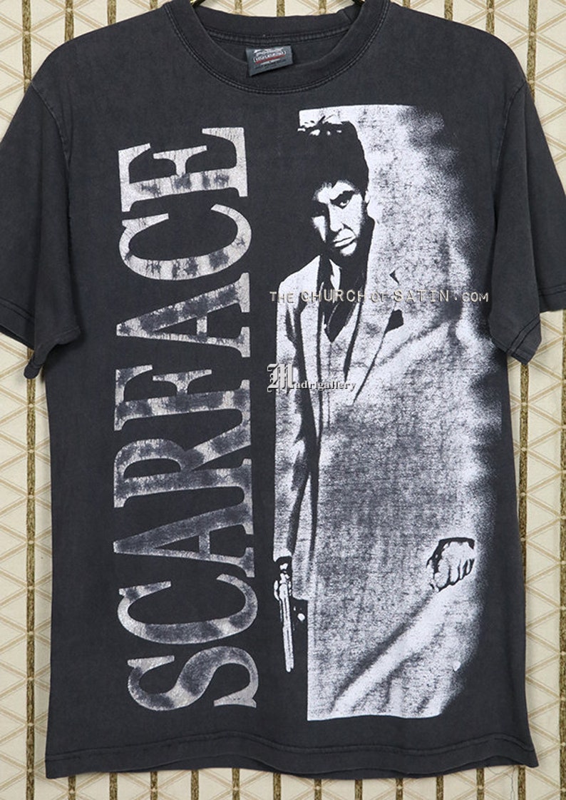 Op de afbeelding: Zwart T-shirt met een witte grafische print van Al Pacino als Tony Montana uit de film Scarface. De tekst "Scarface" is verticaal gedrukt aan de linkerkant van het shirt. De tekst "THE CHURCH OF SATAN.COM" is gedrukt onderaan de grafische print.