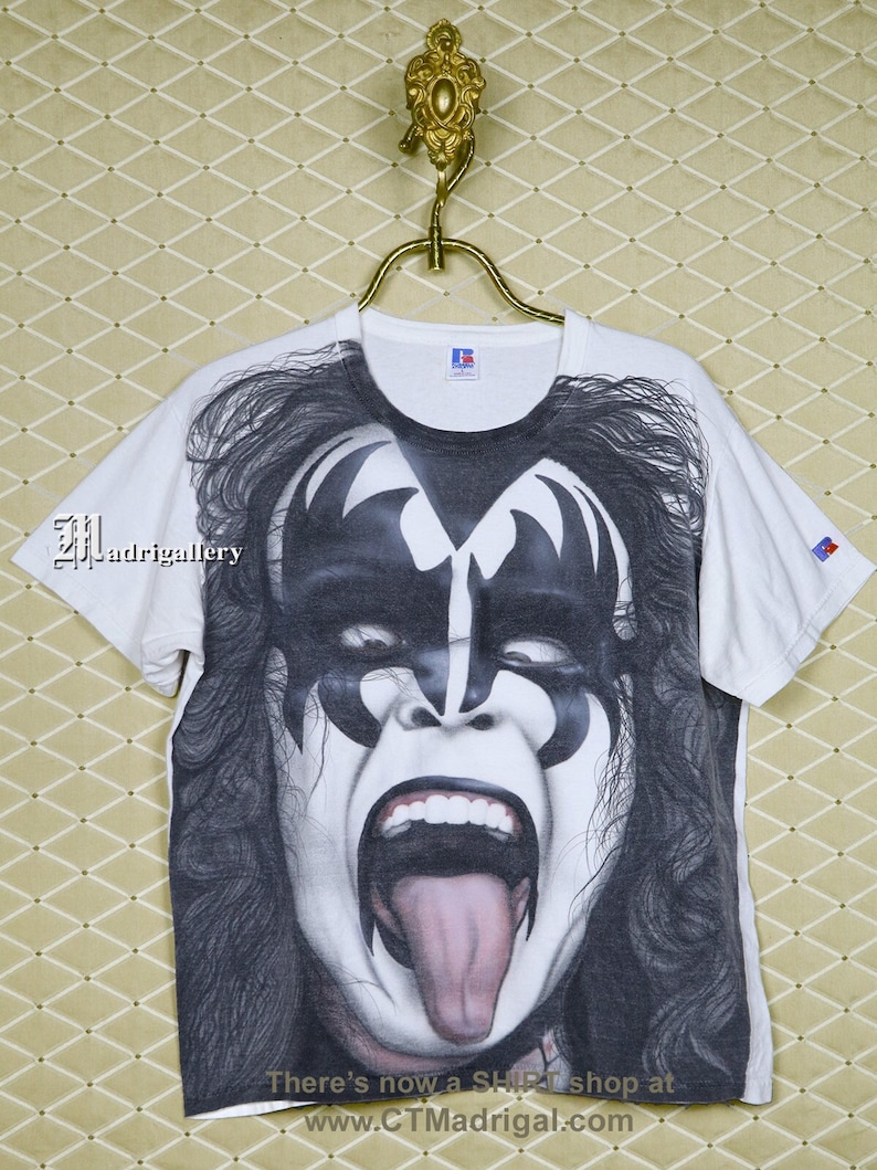 May include: White t-shirt featuring a black and white image of a person with their tongue sticking out. The image is of a rock star with makeup and a black and white design on their face. The text "There's now a Shirt shop at www.CTMadrigal.com" is printed on the bottom of the shirt.