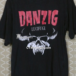 Puede incluir: Camiseta negra con un logotipo rojo de "Danzig" y una calavera blanca con cuernos. El texto "Lucifuge" est&aacute; impreso debajo del logotipo.
