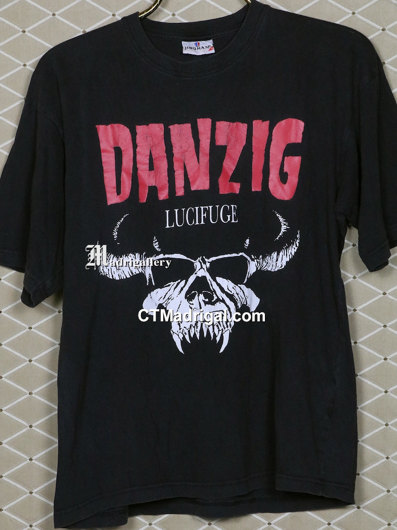 Puede incluir: Camiseta negra con un logotipo rojo de "Danzig" y una calavera blanca con cuernos. El texto "Lucifuge" est&aacute; impreso debajo del logotipo.