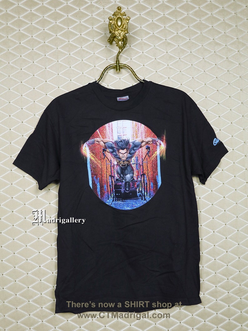 Puede incluir: Camiseta negra con un estampado gr&aacute;fico de un superh&eacute;roe en una escena de ciudad. El gr&aacute;fico est&aacute; en un marco circular con un esquema de color azul, rojo y naranja. El texto "CTMadrigal.com" est&aacute; impreso en la parte inferior de la camiseta.