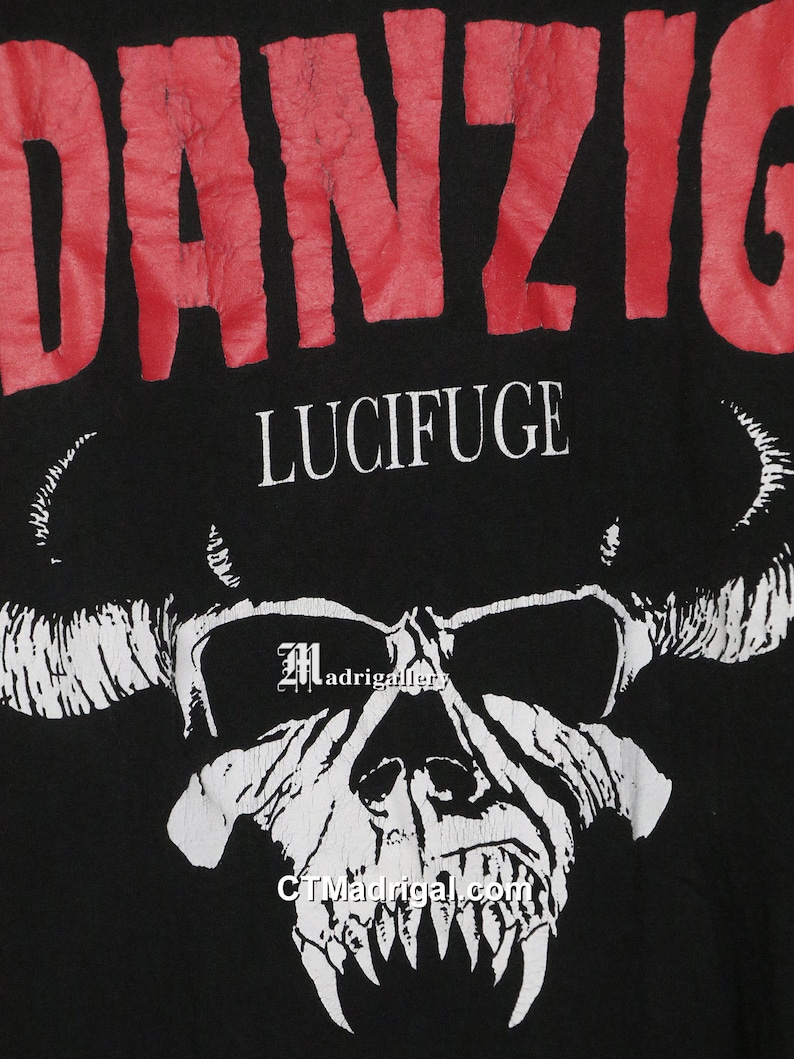 Puede incluir: Camiseta negra con un gr&aacute;fico rojo de "DANZIG" y una calavera blanca con cuernos. El texto "LUCIFUGE" est&aacute; impreso debajo del nombre de la banda.