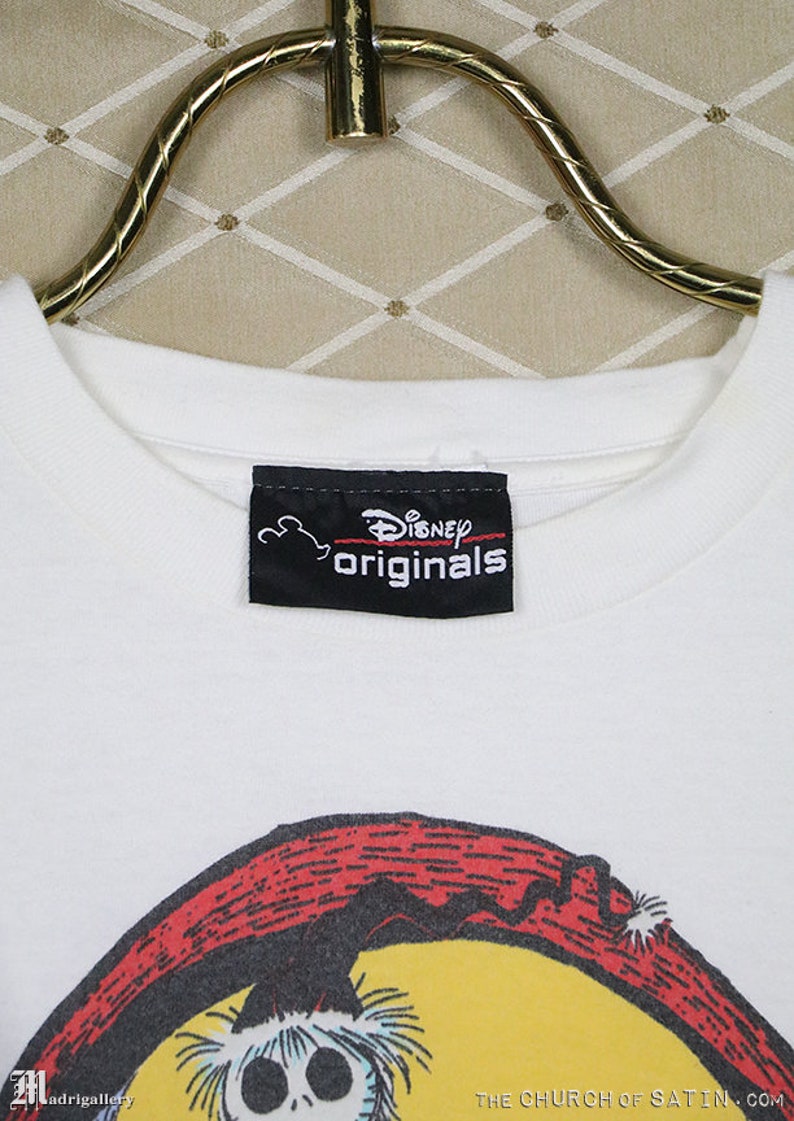 Puede incluir: Camiseta blanca con una etiqueta negra de Disney Originals. La camiseta presenta una ilustraci&oacute;n de dibujos animados de un personaje con una cara de calavera y un fondo rojo y amarillo. El texto "THE CHURCH OF SATIN.COM" est&aacute; impreso en la parte inferior de la camiseta.