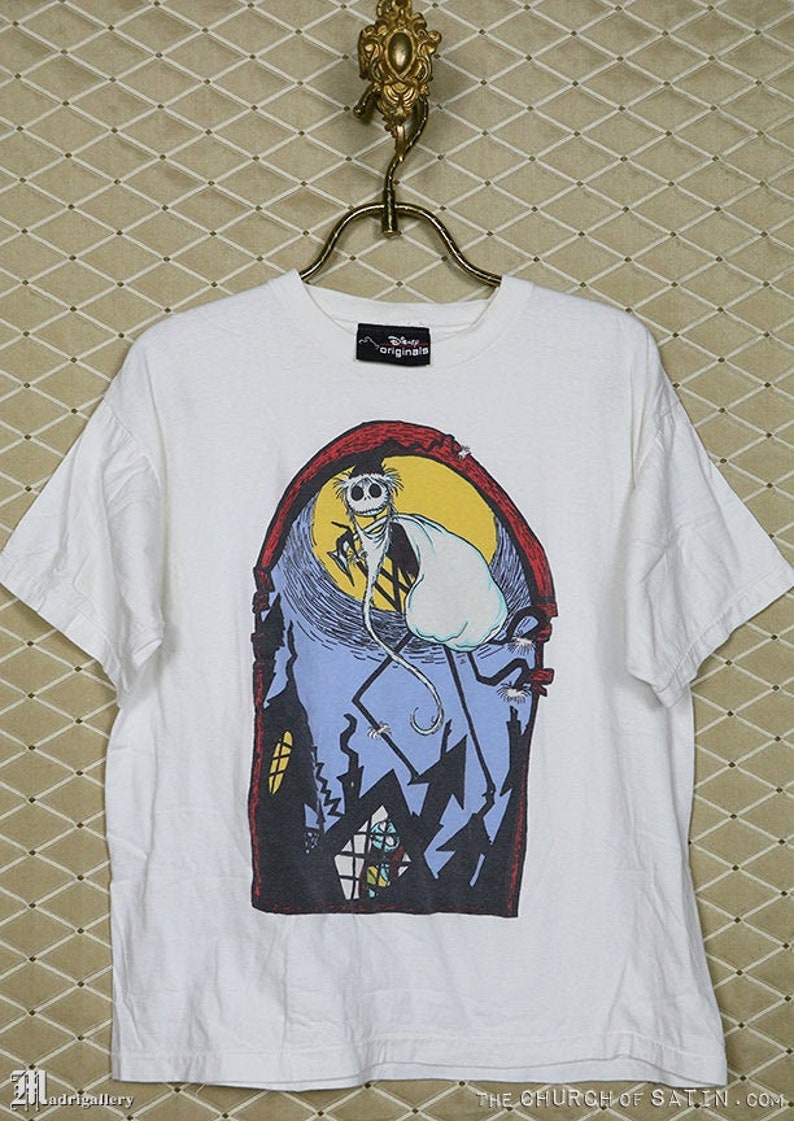 Puede incluir: Camiseta blanca con un estampado gr&aacute;fico de un personaje de dibujos animados de la pel&iacute;cula "Pesadilla antes de Navidad". El personaje est&aacute; de pie en un edificio con una luna llena en el fondo. El texto "Disney Originals" est&aacute; impreso en una etiqueta negra unida a la camiseta.