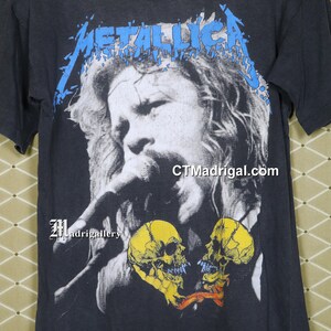 K&ouml;nnte beinhalten: Schwarzes T-Shirt mit einem blauen "Metallica"-Logo und einem Schwarzwei&szlig;bild eines singenden Mannes mit zwei gelben Sch&auml;deln in Herzform. Der Text "CTMadrigal.com" ist in der rechten unteren Ecke des Shirts gedruckt.