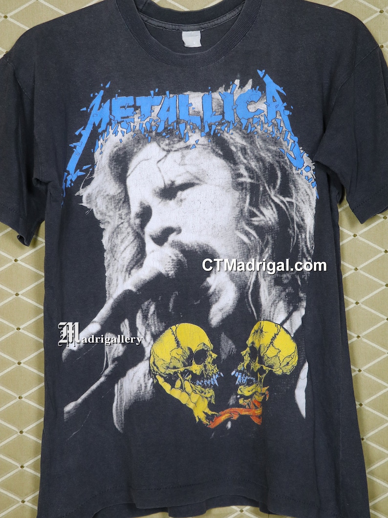 K&ouml;nnte beinhalten: Schwarzes T-Shirt mit einem blauen "Metallica"-Logo und einem Schwarzwei&szlig;bild eines singenden Mannes mit zwei gelben Sch&auml;deln in Herzform. Der Text "CTMadrigal.com" ist in der rechten unteren Ecke des Shirts gedruckt.