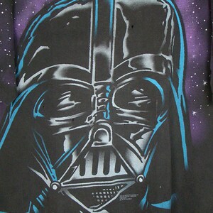 Op de afbeelding: Een zwart T-shirt met een blauw-witte zeefdruk afbeelding van Darth Vaders gezicht tegen een paarse en zwarte galaxy achtergrond.