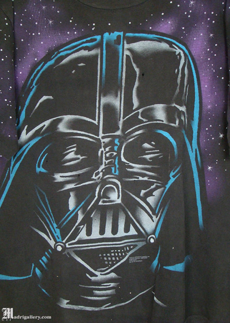 Op de afbeelding: Een zwart T-shirt met een blauw-witte zeefdruk afbeelding van Darth Vaders gezicht tegen een paarse en zwarte galaxy achtergrond.