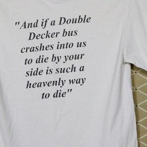 K&ouml;nnte beinhalten: Wei&szlig;es T-Shirt mit schwarzem Text. Der Text lautet: &bdquo;Und wenn uns ein Doppeldeckerbus &uuml;berf&auml;hrt, ist es so himmlisch, an deiner Seite zu sterben.&ldquo; Das Shirt hat auch die Website CTMadrigal.com.