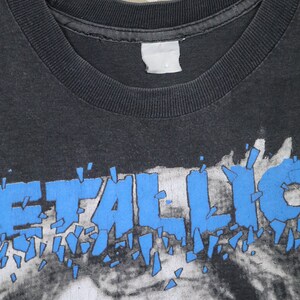 K&ouml;nnte beinhalten: Ein schwarzes T-Shirt mit einem blauen "Metallica"-Logo auf der Vorderseite. Das Logo ist in St&uuml;cke zerbrochen, was einen zersplitterten Effekt erzeugt.