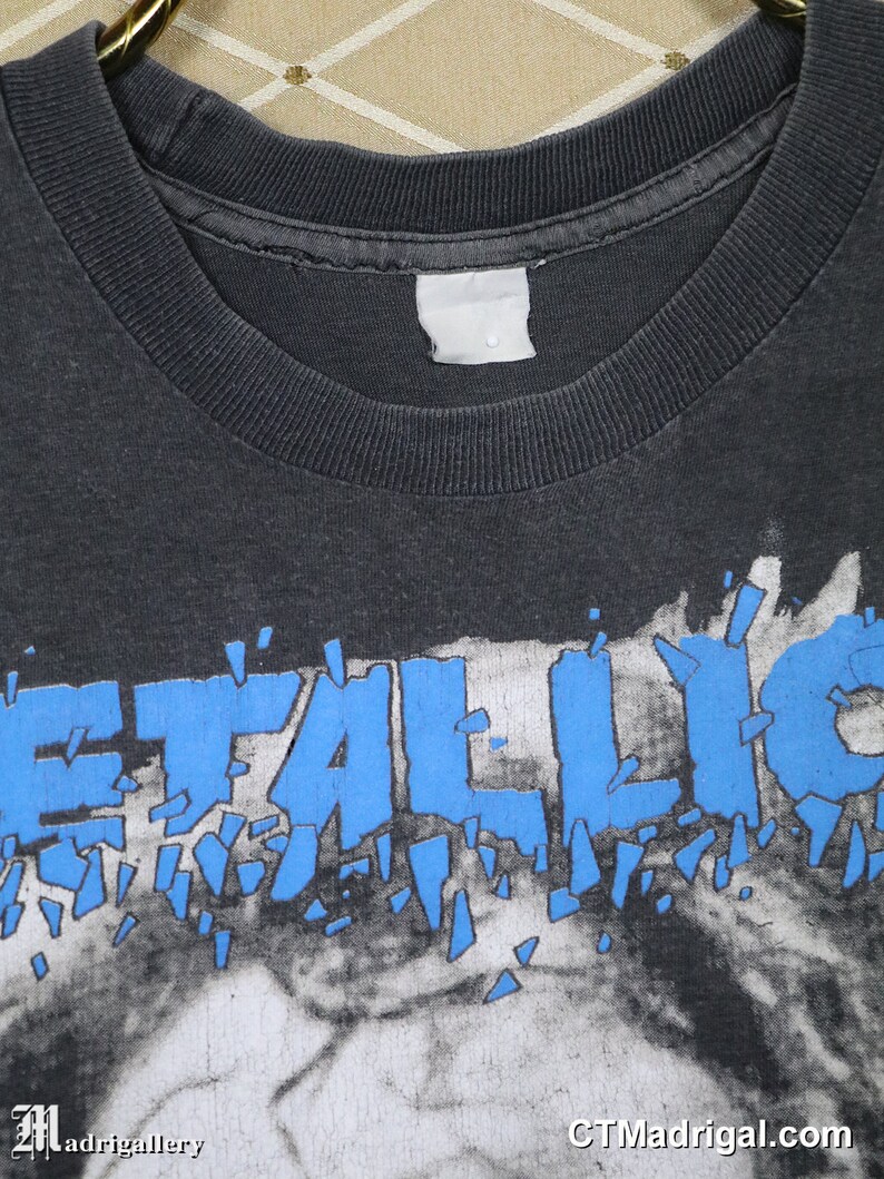 K&ouml;nnte beinhalten: Ein schwarzes T-Shirt mit einem blauen "Metallica"-Logo auf der Vorderseite. Das Logo ist in St&uuml;cke zerbrochen, was einen zersplitterten Effekt erzeugt.