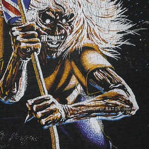 May include: A black t-shirt with a graphic print of a metal band mascot. The mascot is a monstrous figure with long white hair, holding a flag with a red, white, and blue design. The text "THE CHURCH OF SATIN.COM" is printed on the shirt.