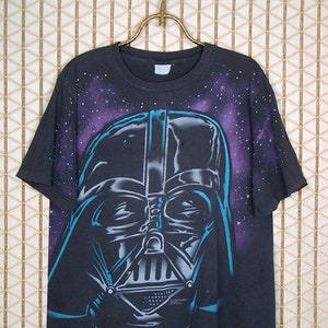 Op de afbeelding: Zwart T-shirt met een grote, kleurrijke afbeelding van Darth Vaders gezicht tegen een paarse en blauwe galactische achtergrond.