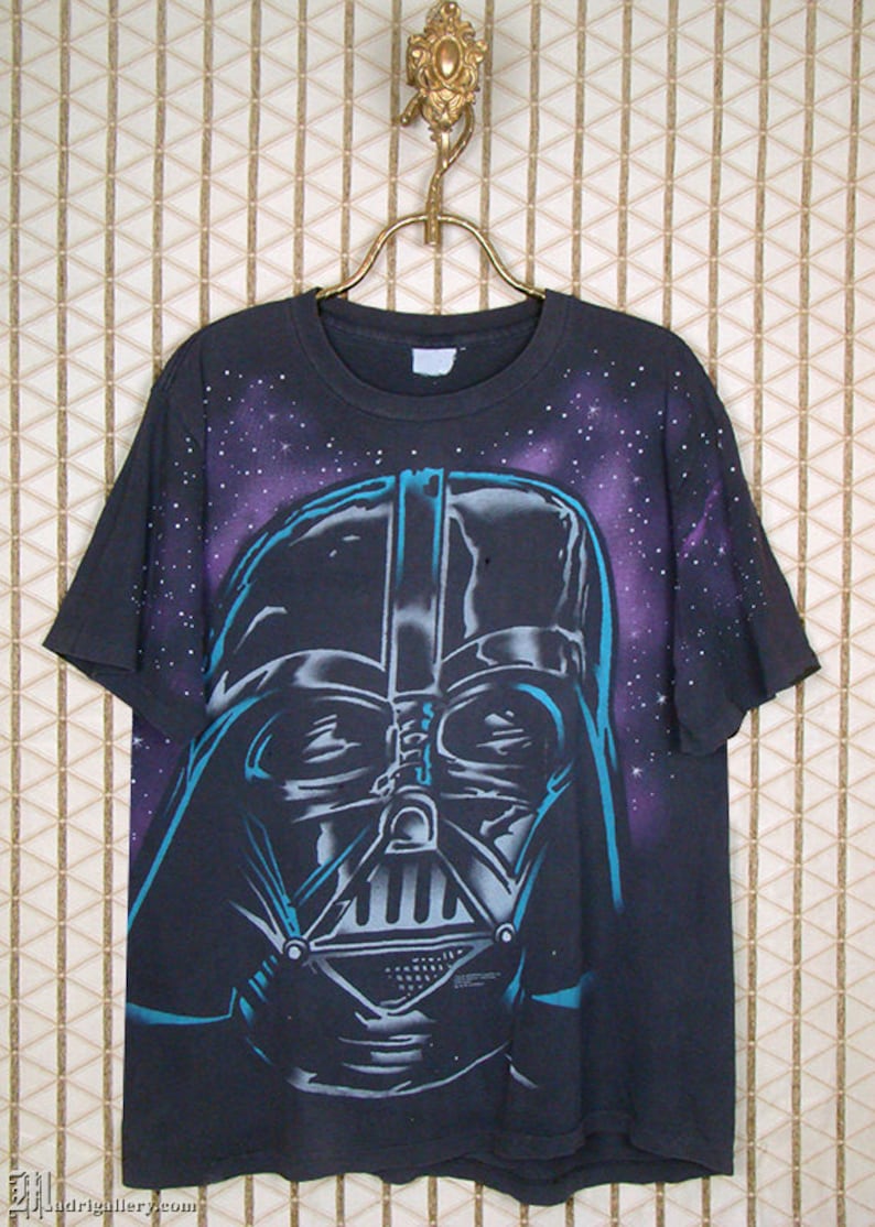 Op de afbeelding: Zwart T-shirt met een grote, kleurrijke afbeelding van Darth Vaders gezicht tegen een paarse en blauwe galactische achtergrond.