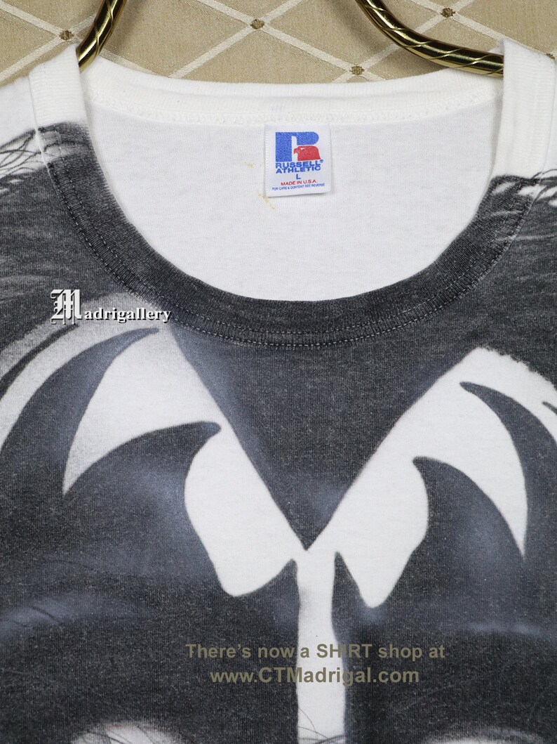 May include: White t-shirt with a black and white graphic print of a skull with wings. The shirt is made by Russell Athletic and is size L. The text "There's now a SHIRT shop at www.CTMadrigal.com" is printed on the bottom of the shirt.
