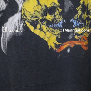 K&ouml;nnte beinhalten: Schwarzes T-Shirt mit einem grafischen Druck von zwei gelben Sch&auml;deln mit blauen Z&auml;hnen. Die Sch&auml;del sind einander zugewandt und haben ein rotes und orangefarbenes Band zwischen sich. Der Text "CTMadrigal.com" ist unten rechts auf dem T-Shirt aufgedruckt.