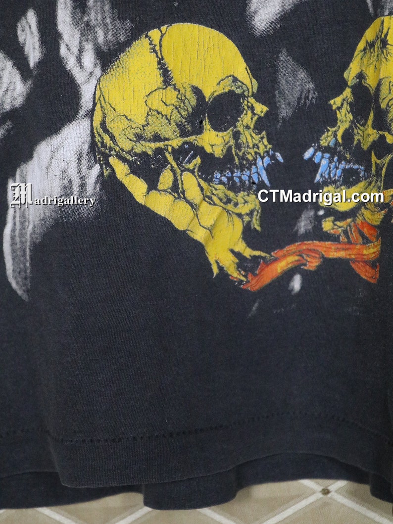 K&ouml;nnte beinhalten: Schwarzes T-Shirt mit einem grafischen Druck von zwei gelben Sch&auml;deln mit blauen Z&auml;hnen. Die Sch&auml;del sind einander zugewandt und haben ein rotes und orangefarbenes Band zwischen sich. Der Text "CTMadrigal.com" ist unten rechts auf dem T-Shirt aufgedruckt.